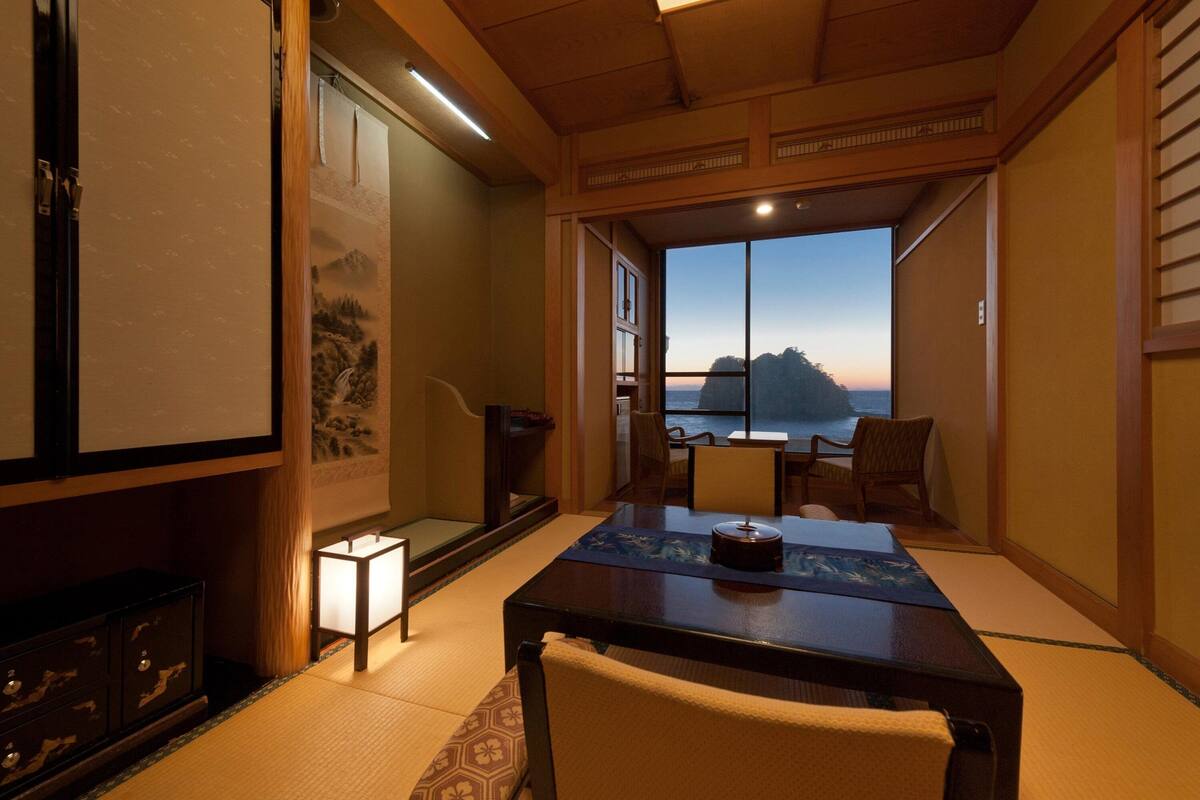 Una habitación japonesa tradicional con suelo de tatami, una mesa baja con un juego de té y vistas a un paisaje montañoso a través de un ventanal grande.
