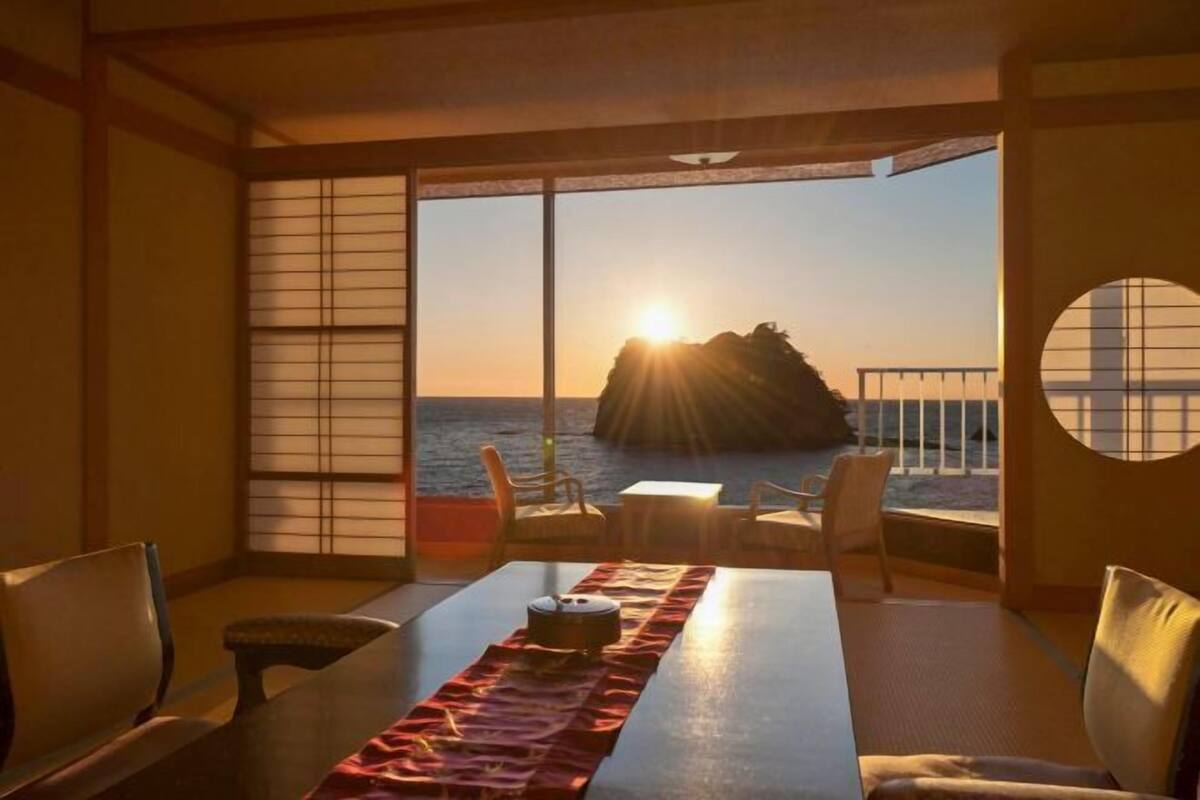 Una habitación con suelo de tatami, una mesa baja con un tapete rojo, sillas y vista al mar al atardecer.