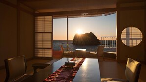 Una habitación japonesa tradicional con suelo de tatami, una mesa baja con un tapete rojo y vistas al mar al atardecer.