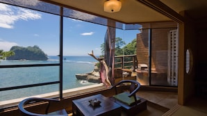 Una habitación con un amplio ventanal con vistas a un litoral rocoso y al océano. Hay una mesa de madera con una silla, un pequeño adorno sobre la mesa y un tapete tradicional japonés en el suelo.