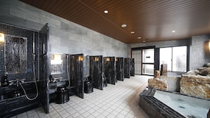 A modern bathroom with multiple shower stalls, a large mirror, and a seating area with a water feature. - Hotel Dormy Inn Miyazaki Natural Hot Spring (Miyazaki)