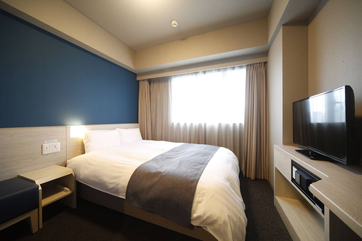 A hotel room with a bed, bedside table, TV, and a window with curtains.