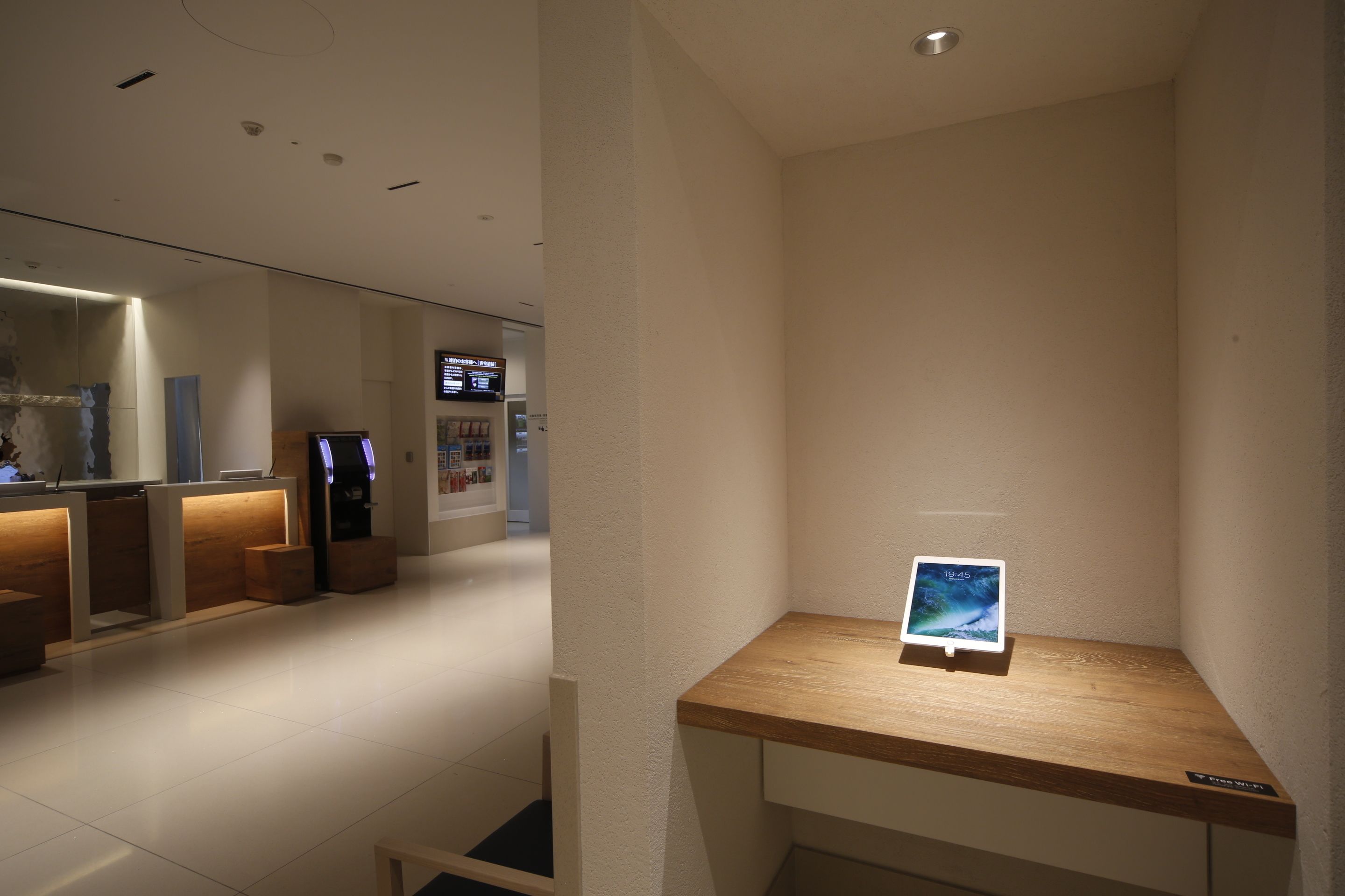 A modern hotel lobby with a reception desk, a digital information kiosk, and a wooden display table with a tablet.