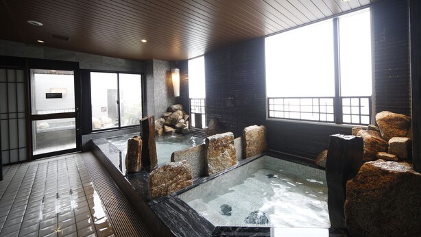 A traditional Japanese onsen with a wooden ceiling, stone seating, and a view of the city through large windows. - Hotel Dormy Inn Miyazaki Natural Hot Spring (Miyazaki)