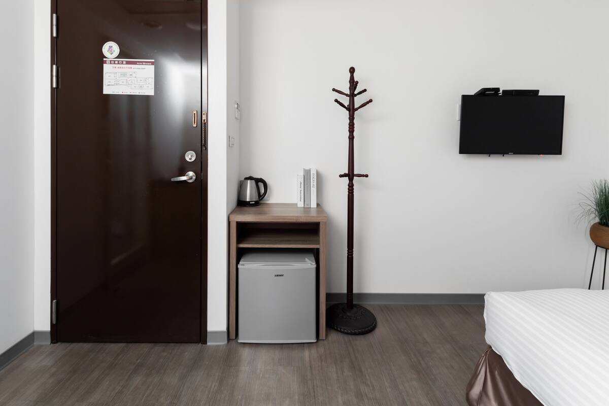 A hotel room with a closed door, a small refrigerator, a coat rack, a flat-screen TV, and a bed with a white bedspread.