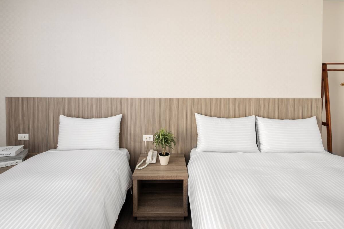 A hotel room with two beds, a wooden headboard, a bedside table with a phone and a plant, and a small shelf with books.