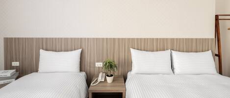 A hotel room with two beds, a wooden headboard, a bedside table with a phone and a plant, and a small shelf with books.