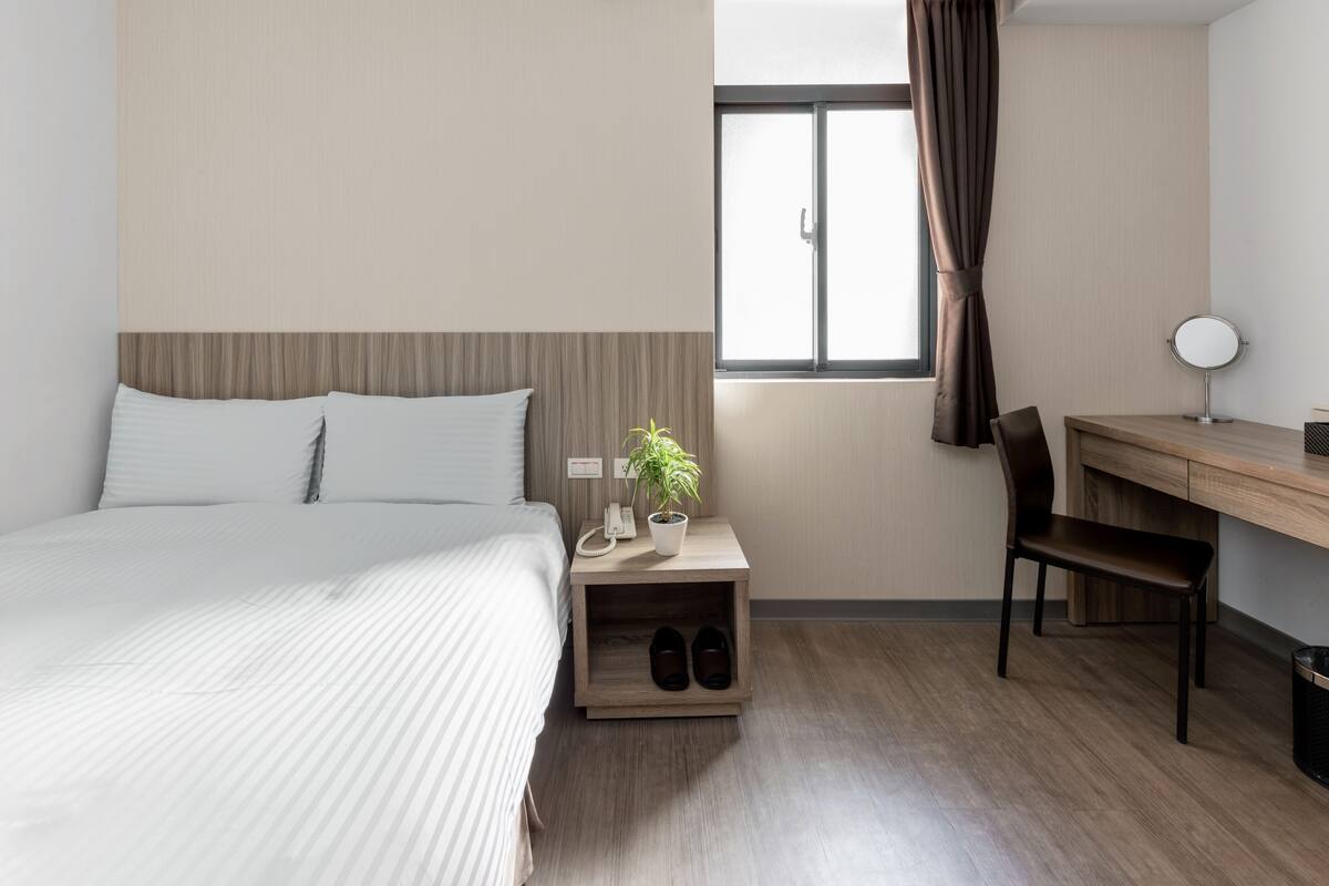A hotel room with a bed, a desk, a chair, a potted plant, a mirror, and a window with curtains.