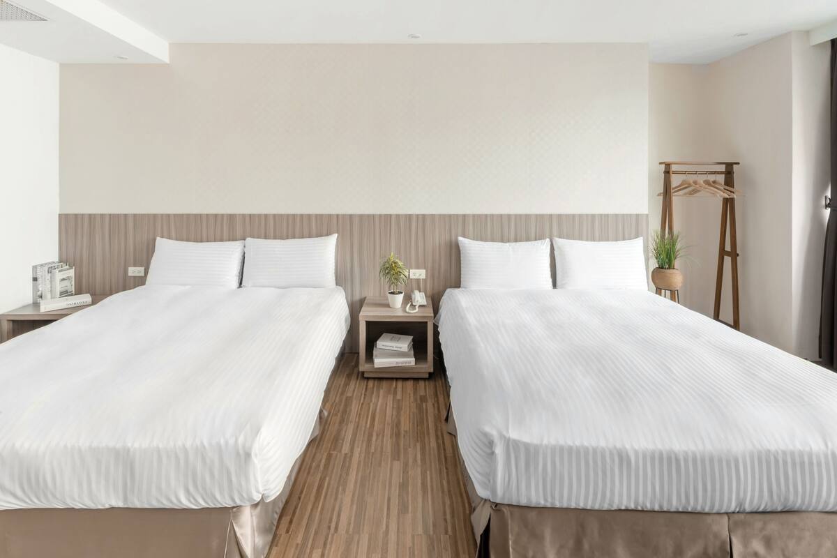 A hotel room with two beds, a wooden headboard, a bedside table with a lamp, a small plant, and a coat rack.