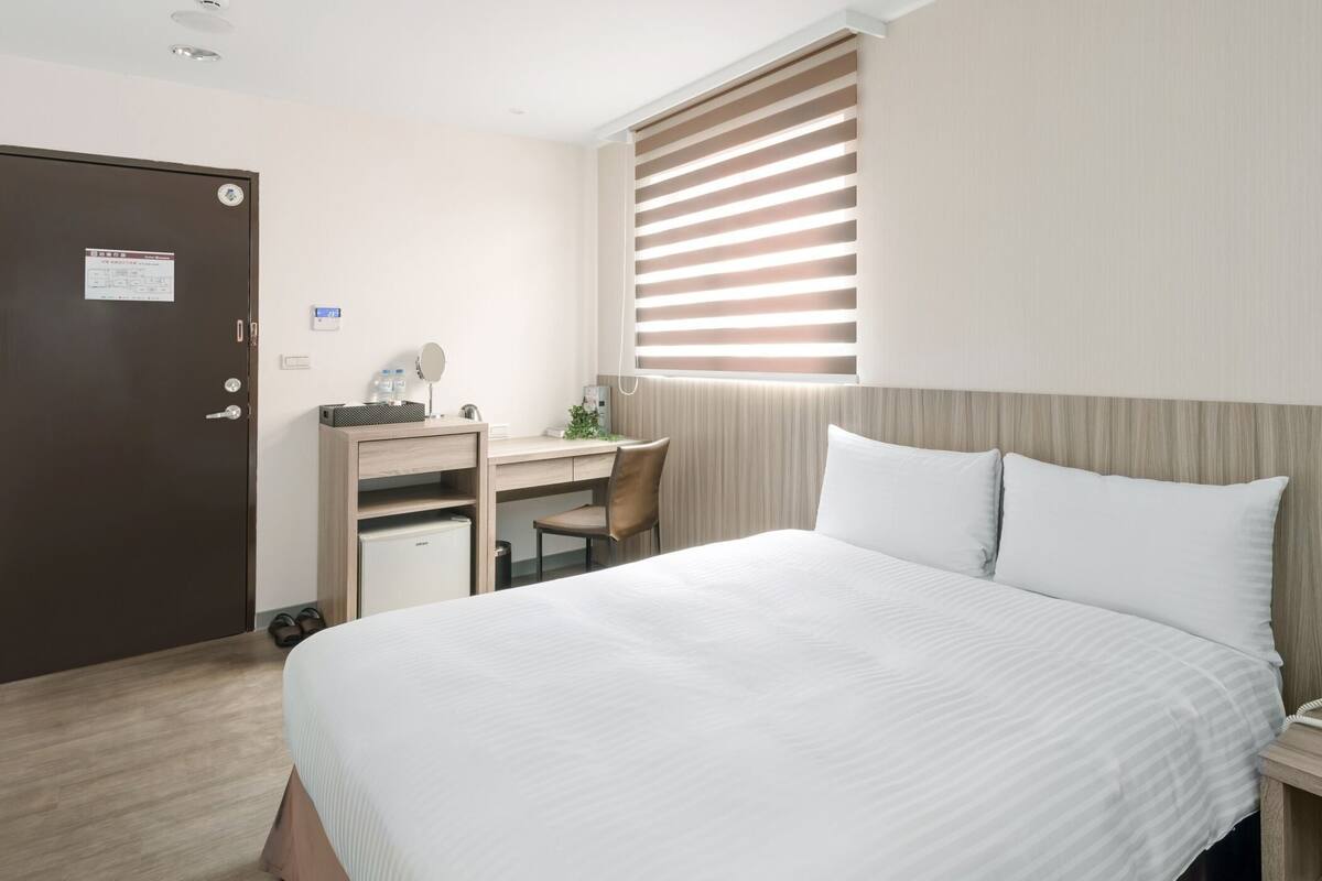 A hotel room with a bed, a desk, a chair, a small fridge, a phone, and a window with blinds.