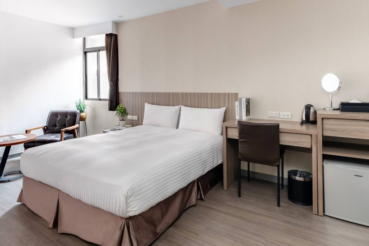 A hotel room with a large bed, a desk with a chair, a small table, a plant, a window with curtains, and a minibar.