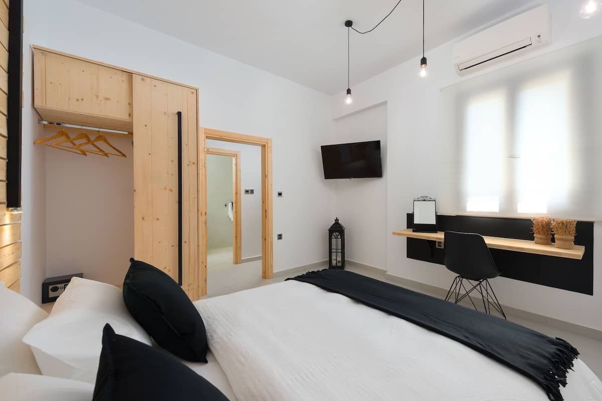 A modern bedroom with a bed, a wooden wardrobe, a desk with a chair, a TV, and a window with natural light.