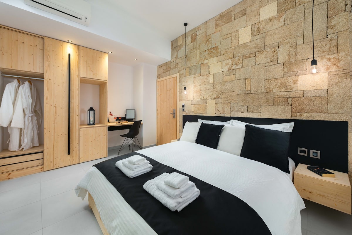 A modern bedroom with a stone wall, a wooden wardrobe, a desk, and a neatly made bed.