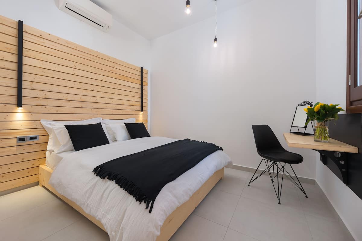 A modern bedroom with a wooden headboard, a bed with white and black bedding, a black chair, and a small desk with a vase of flowers.
