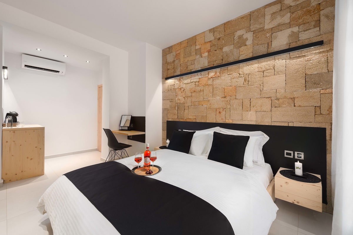 A modern hotel room with a stone wall, a bed with black and white bedding, a wooden nightstand, and a desk with a chair.