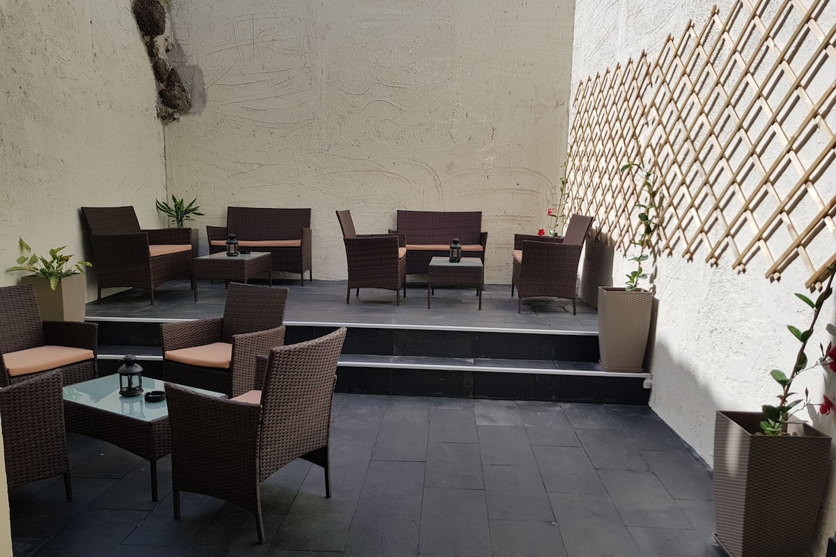 An outdoor patio area with wicker furniture, a glass table, and potted plants.
