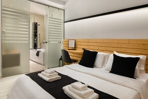 Una habitación de hotel moderna con una cama grande, cabecera de madera, un escritorio con silla y cabina de ducha de vidrio.