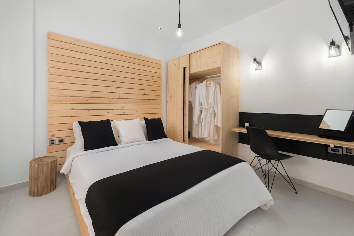 A modern bedroom with a wooden headboard, a bed with black and white bedding, a desk with a chair, and a walk-in closet with hanging clothes.