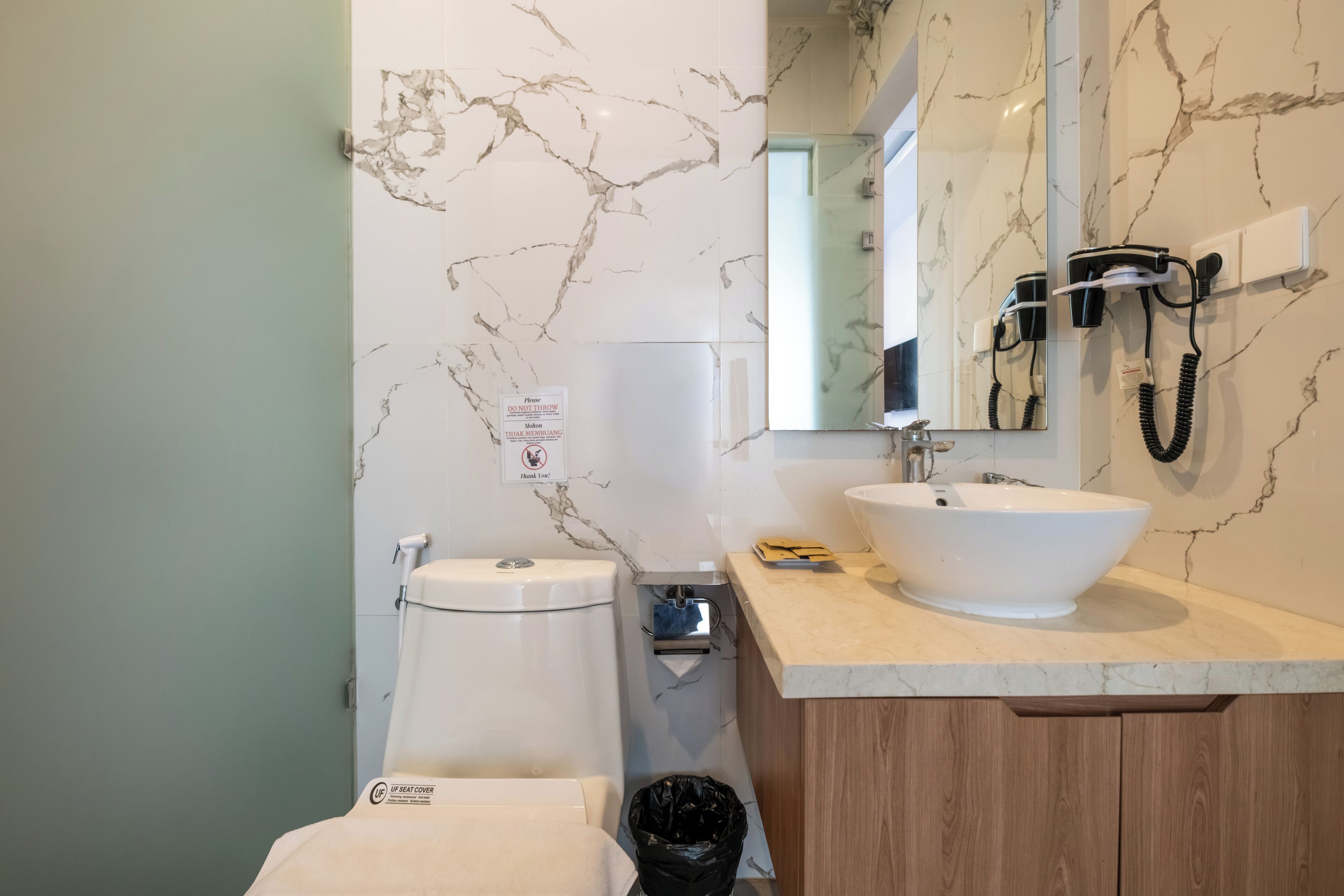 Een badkamer met een marmeren wand, een toilet, een wastafel, een haardroger en een vuilnisbak.