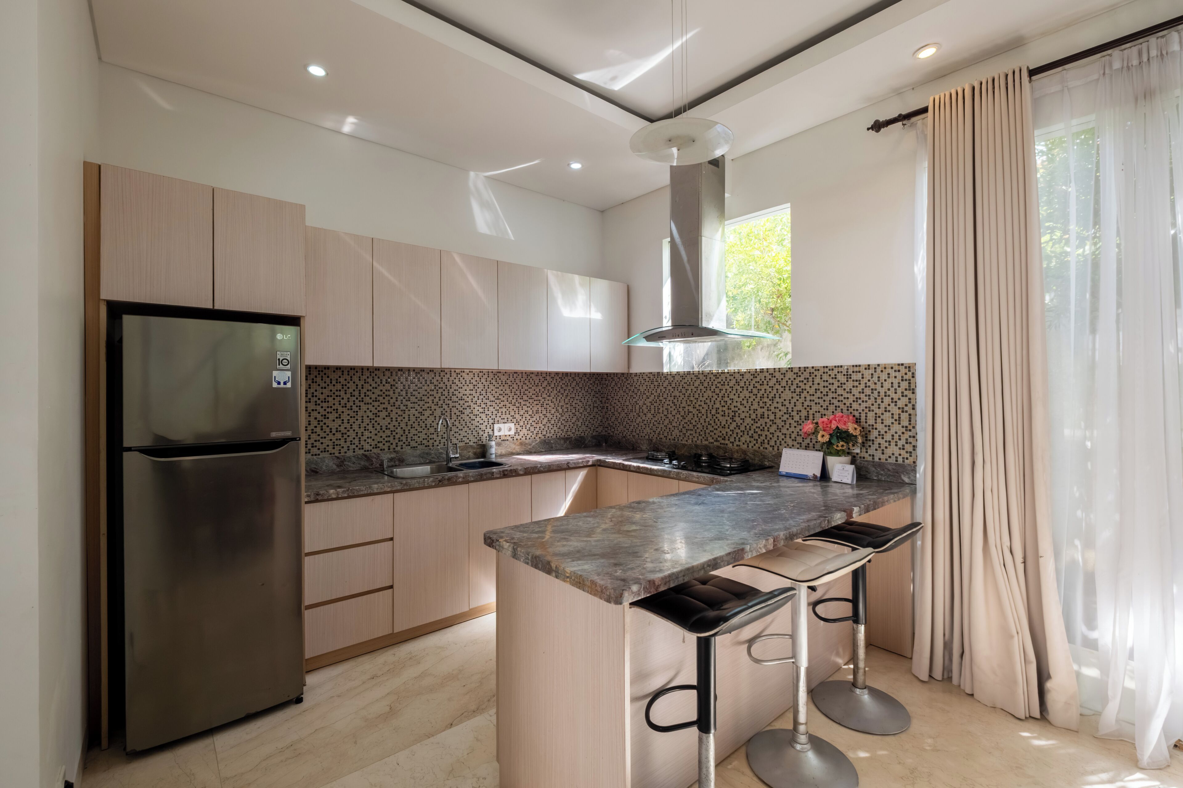 A modern kitchen with a built-in island, stainless steel refrigerator, and a window with curtains.