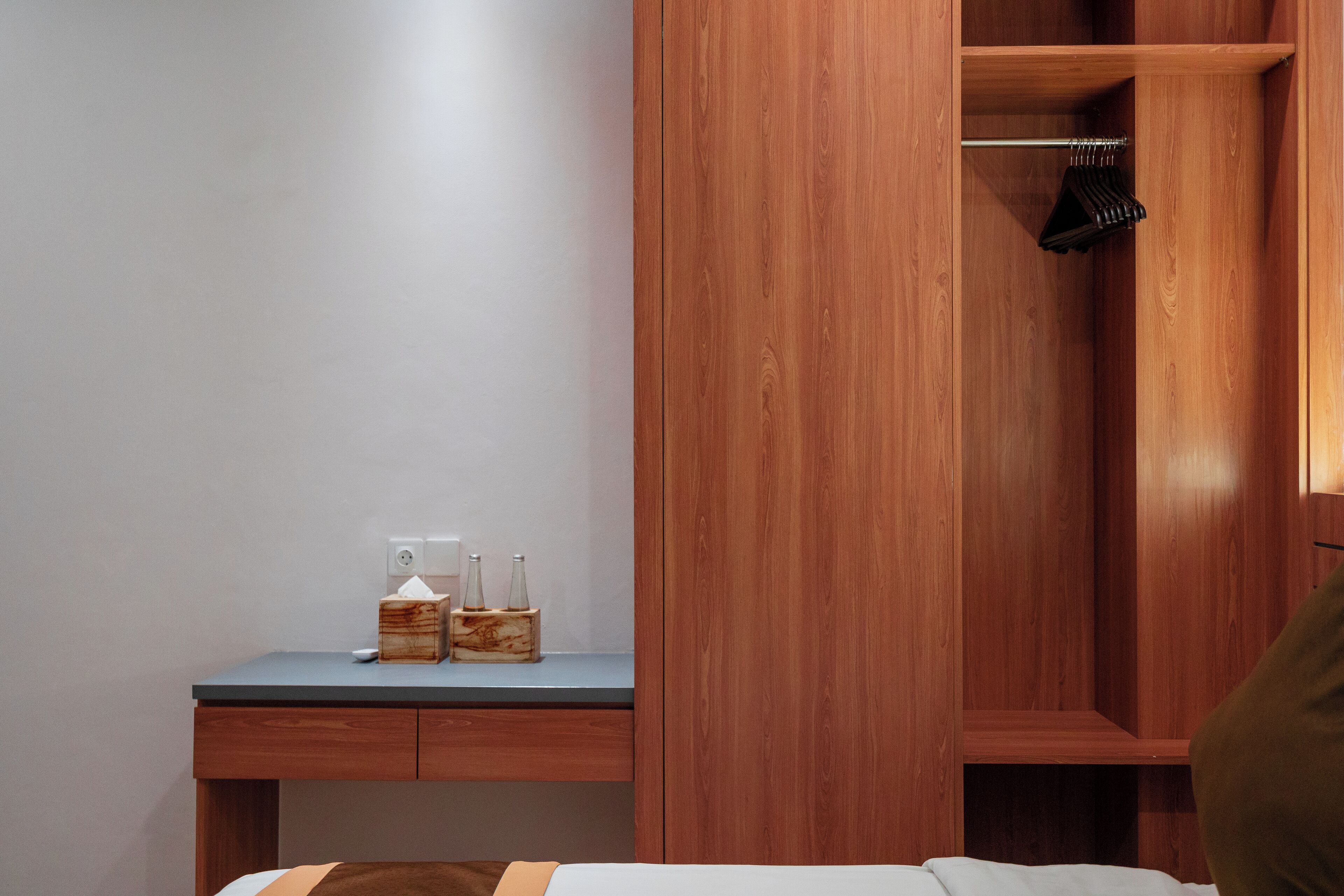 A bedroom with a wooden wardrobe, a bedside table with a tissue box and bottles, and a hanging coat rack.