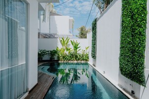 A modern swimming pool with a wooden deck, surrounded by white walls and lush greenery.