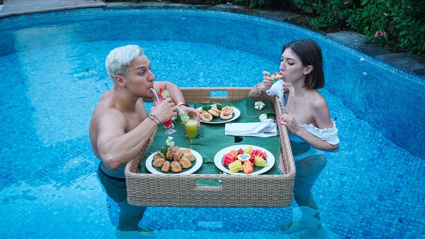 Two people dining by a poolside with a wicker table holding plates of food and drinks. - Bali Residence Villa (Ungasan)