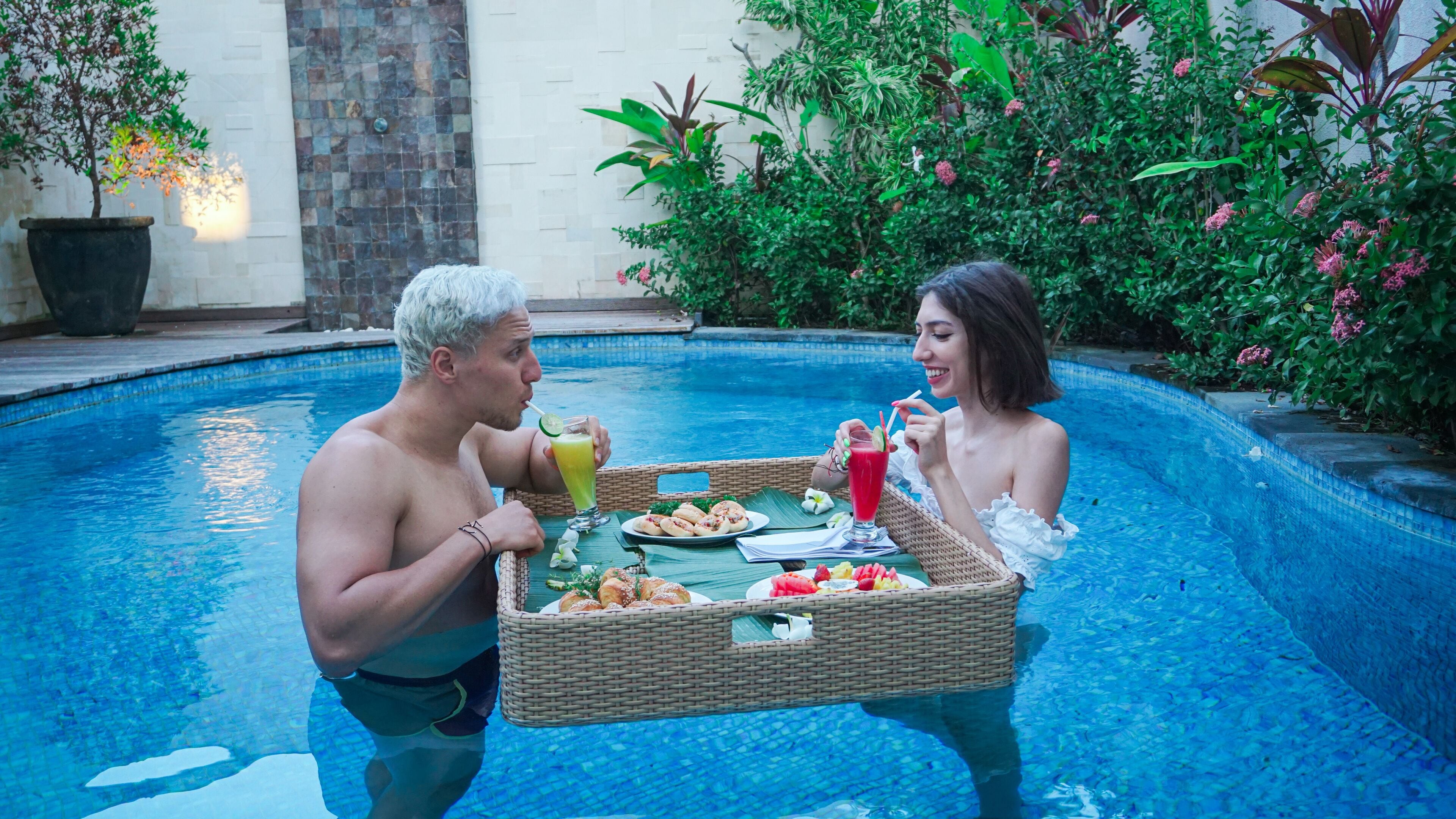 Two people by a pool with a wicker table holding food and drinks.