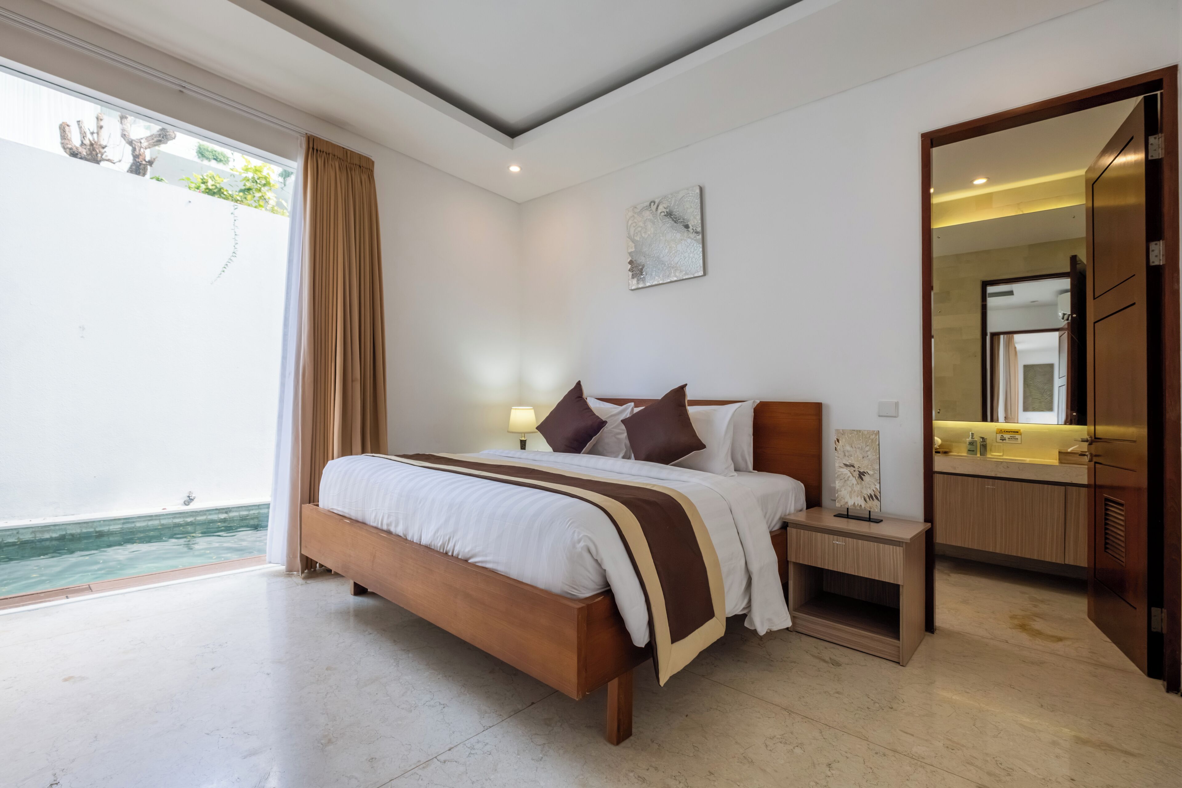 A modern bedroom with a large bed, bedside tables, a mirror, and a view of a pool.