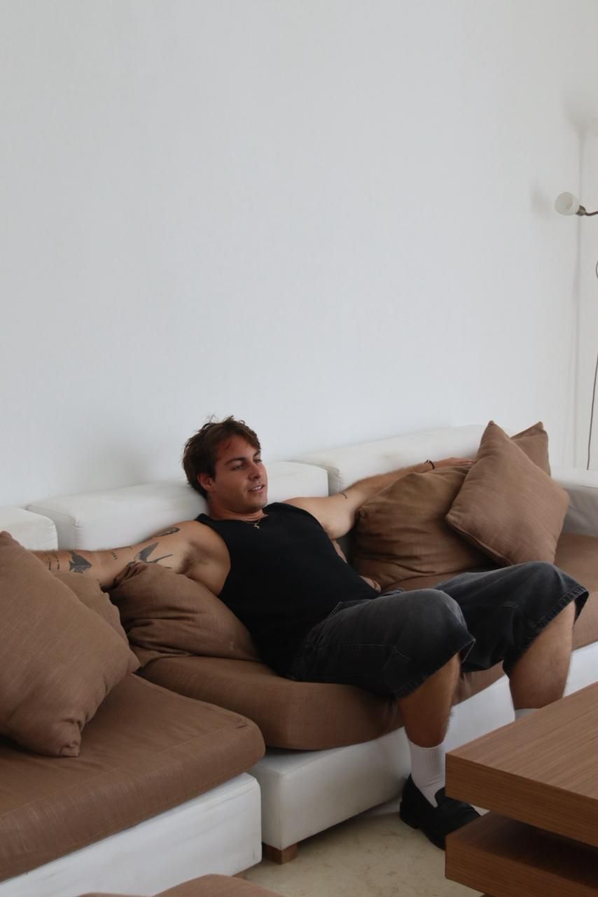 Een persoon ligt op een witte bank met bruine kussens in een kamer met een witte muur en een houten tafel.