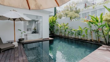 Un espace piscine extérieur moderne, doté d’une terrasse en bois, d’une chaise longue et d’un grand parasol offrant de l’ombre.