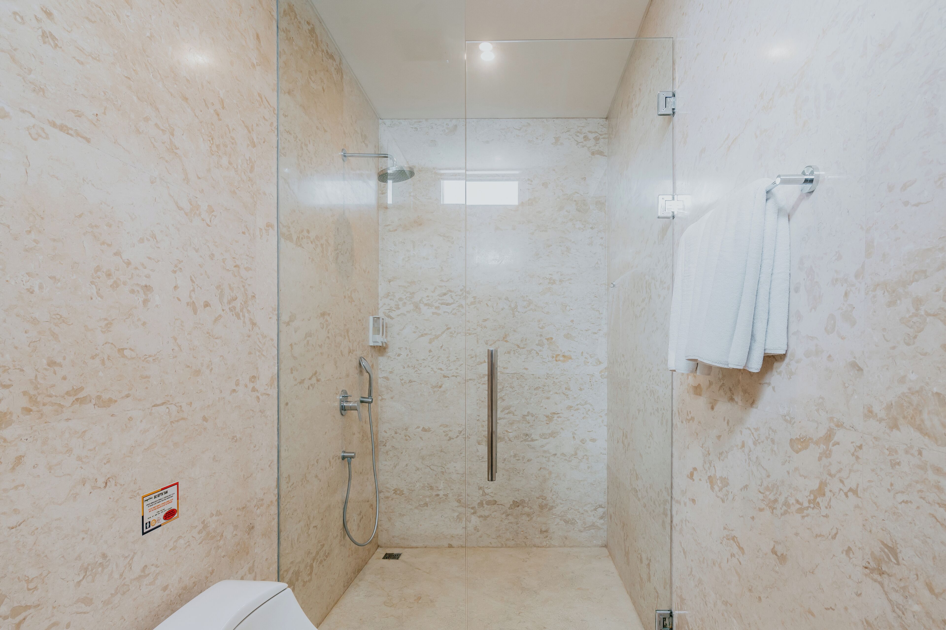 A modern bathroom with a glass-enclosed shower, a white towel rack, and a visible warning sign on the wall.