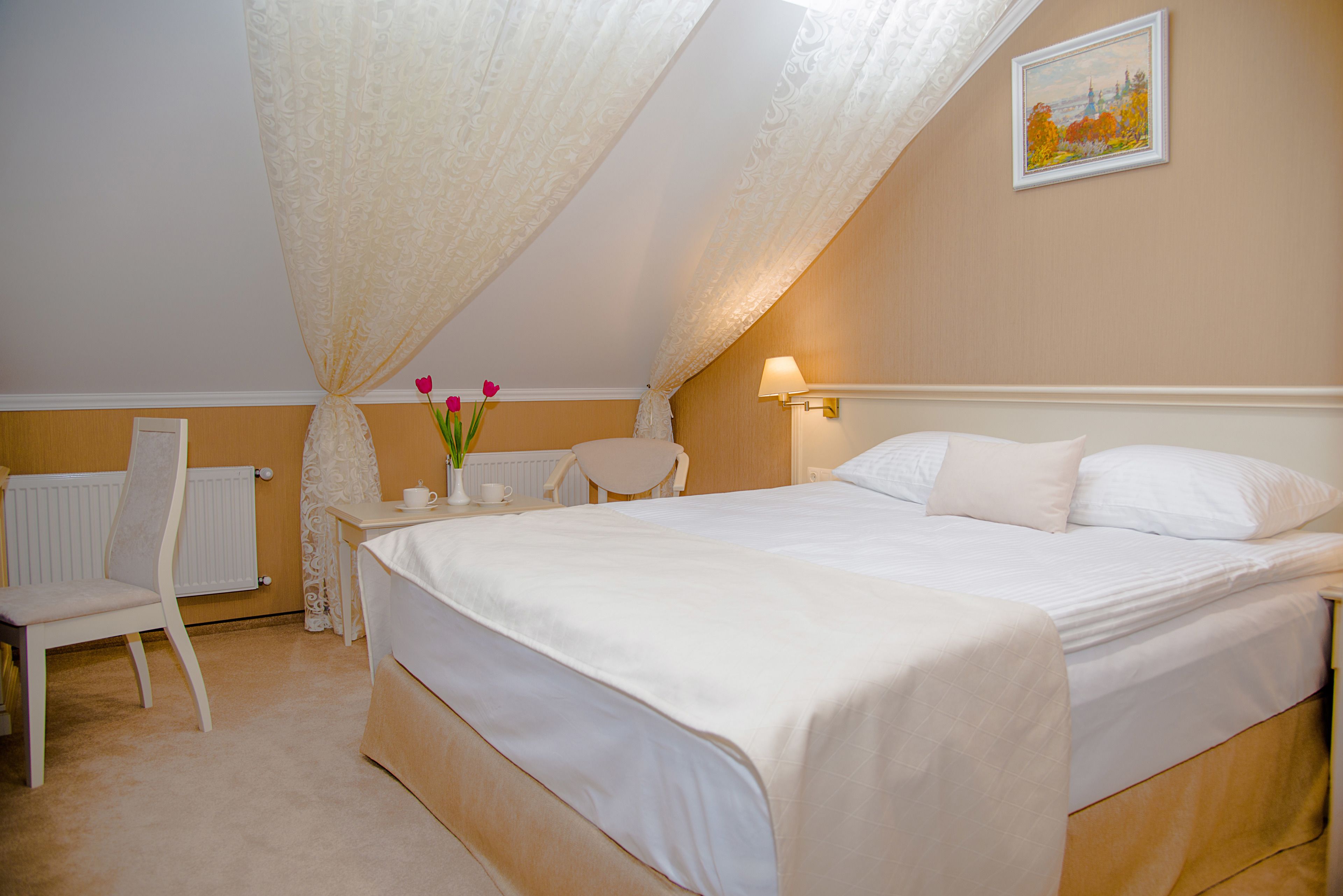 Ein Schlafzimmer mit einem großen Bett, einem Sessel, einem Tisch mit Blumenvase, einem Heizkörper und einem Bild an der Wand.