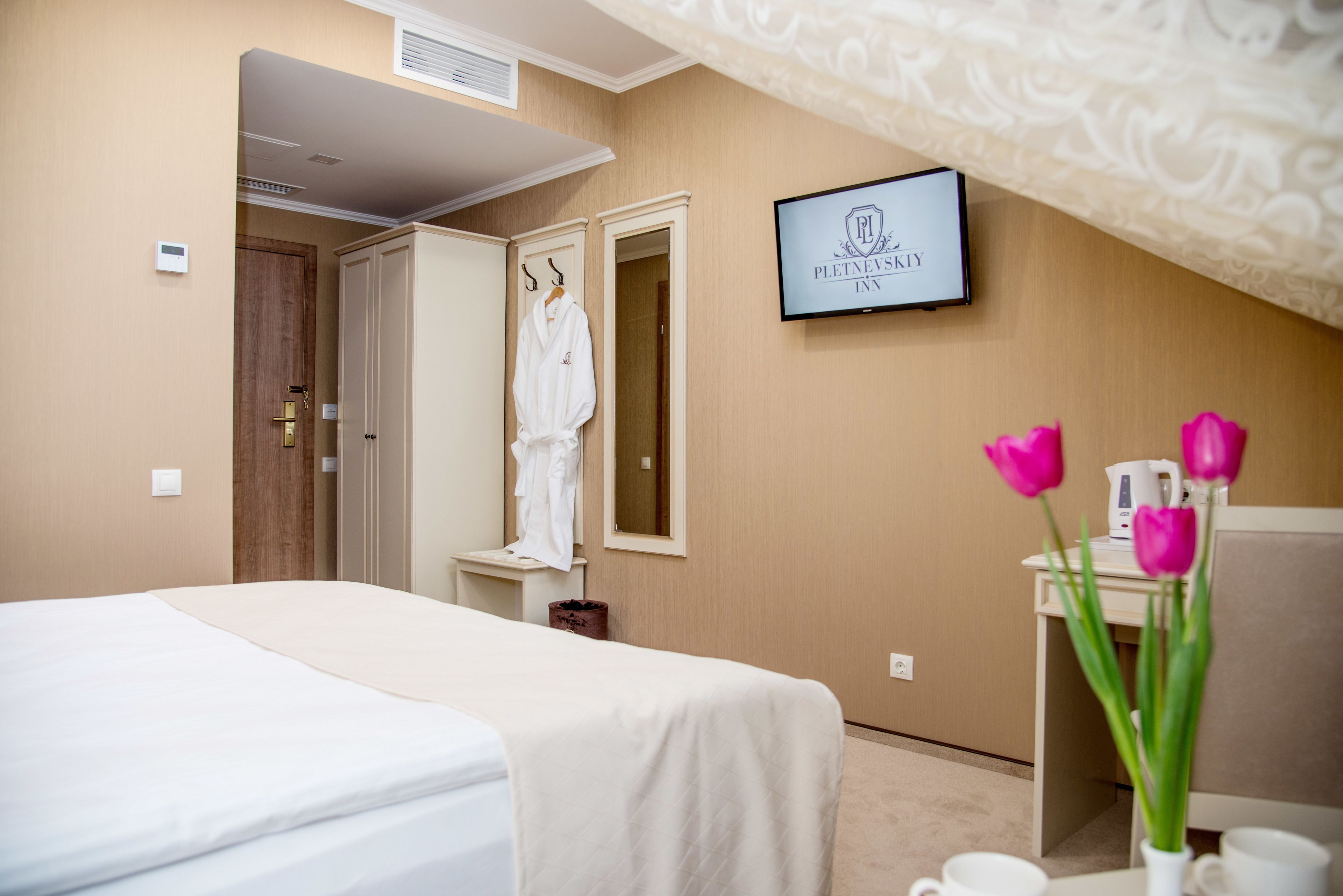 Une chambre d’hôtel avec un lit, un peignoir accroché à la porte, un téléviseur fixé au mur et un vase rempli de fleurs.