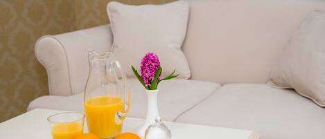 Una mesa blanca con una jarra de vidrio con jugo de naranja, dos vasos de jugo de naranja y dos naranjas. También hay un florero de vidrio con flores rosas sobre la mesa.