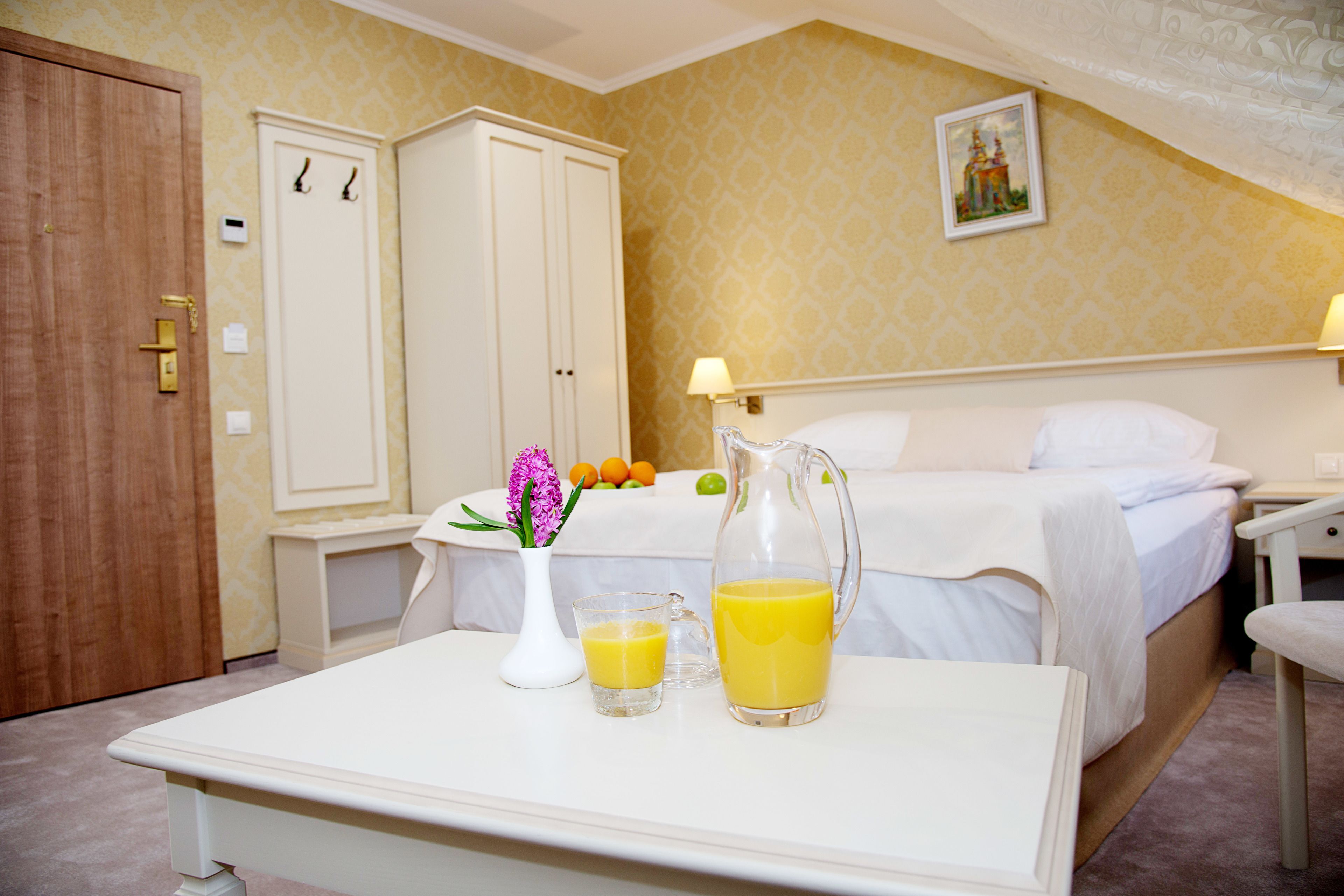 Een hotelkamer met een bed, een nachtkastje met een karaf sinaasappelsap, een vaas met bloemen en een tafeltje met een glas sinaasappelsap.
