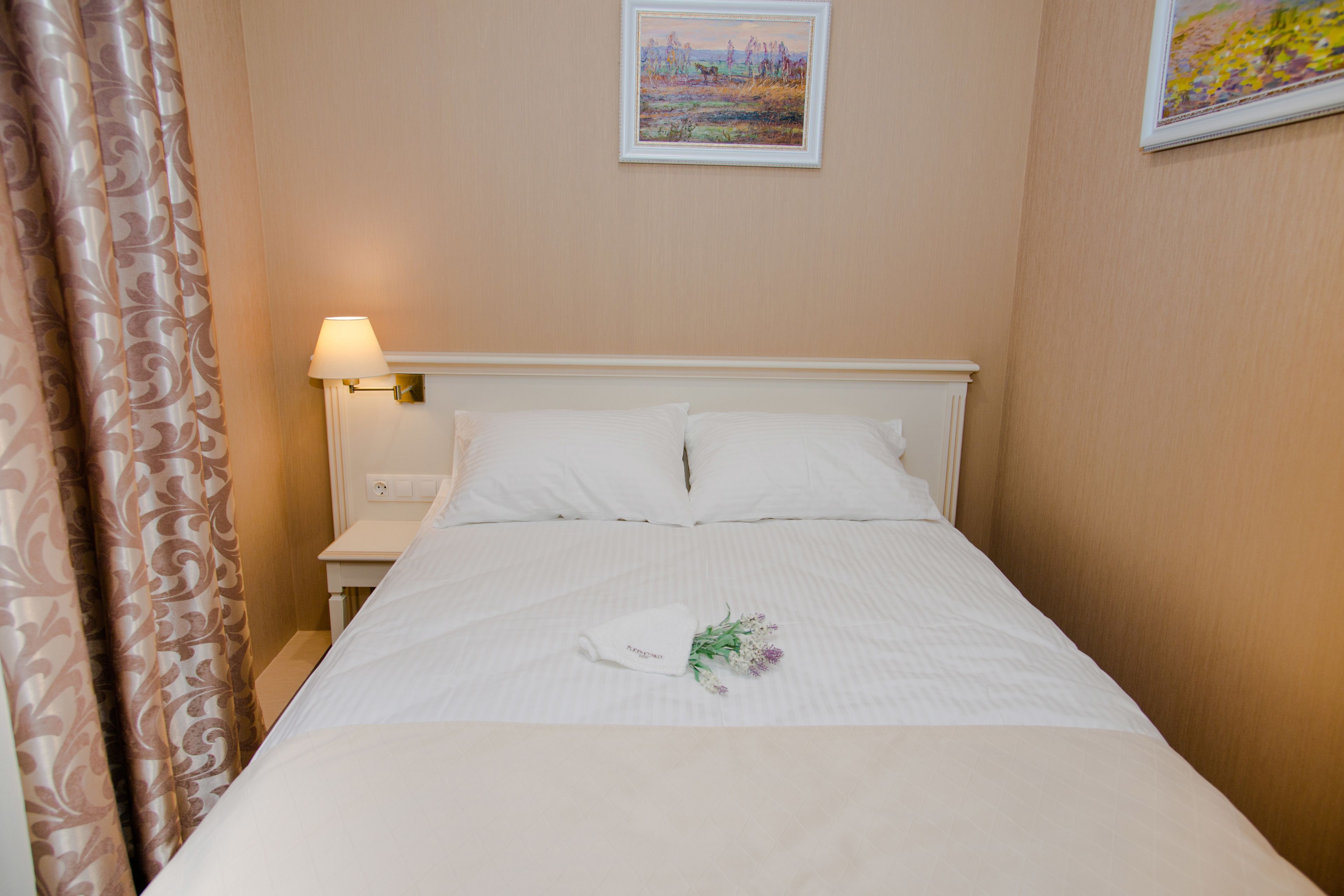 A neatly made bed with white linens and a floral arrangement, a bedside lamp, and a painting on the wall.