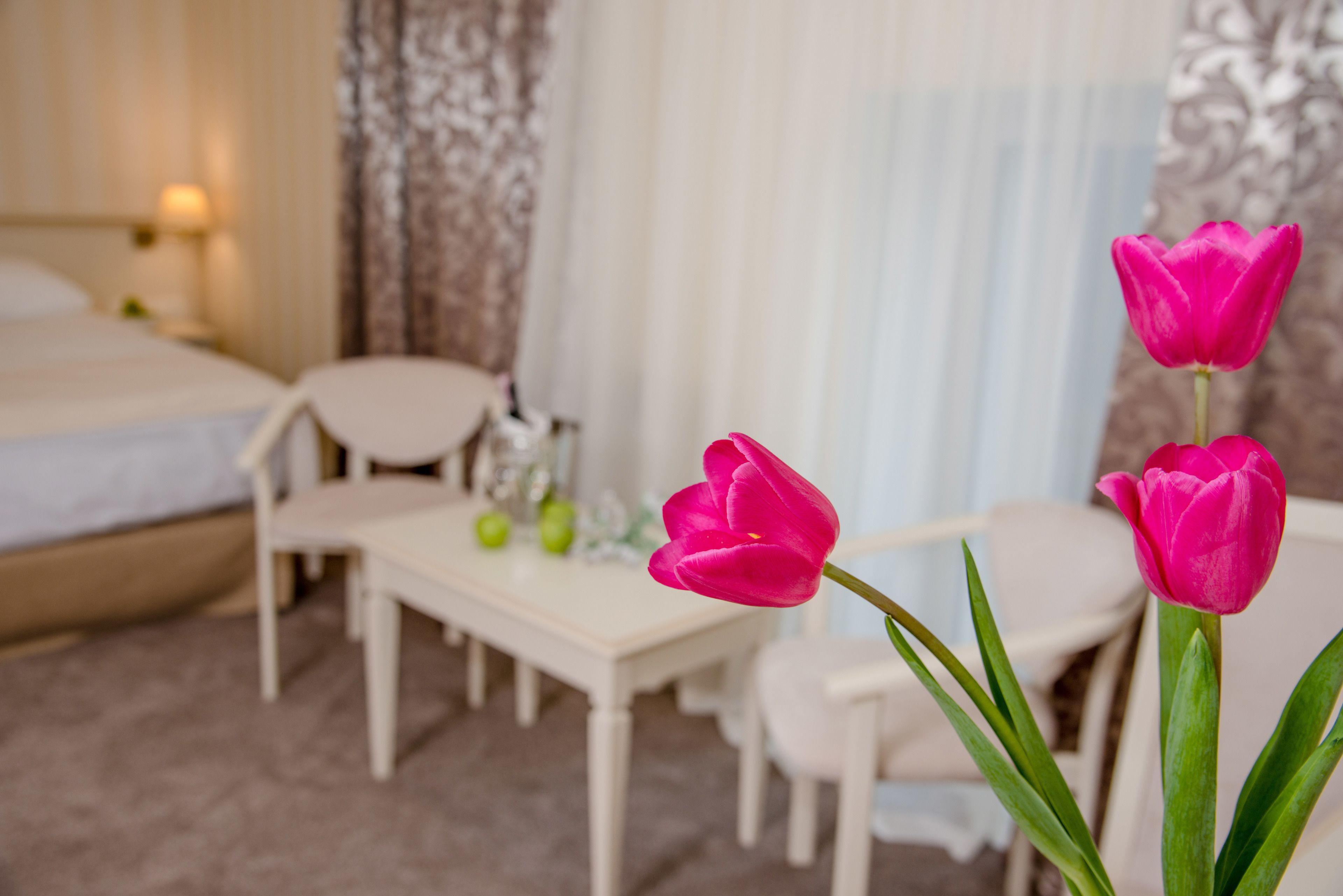 A bedroom with a bed, a small table, and two chairs. There are two pink tulips in the foreground.