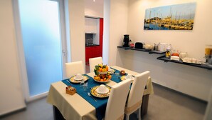 Een eethoek met een tafel voor vier personen, een keuken op de achtergrond en een schilderij van een jachthaven aan de muur.