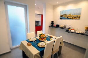 Zona pranzo con tavolo apparecchiato per quattro, cucina sullo sfondo e un quadro raffigurante un porto.