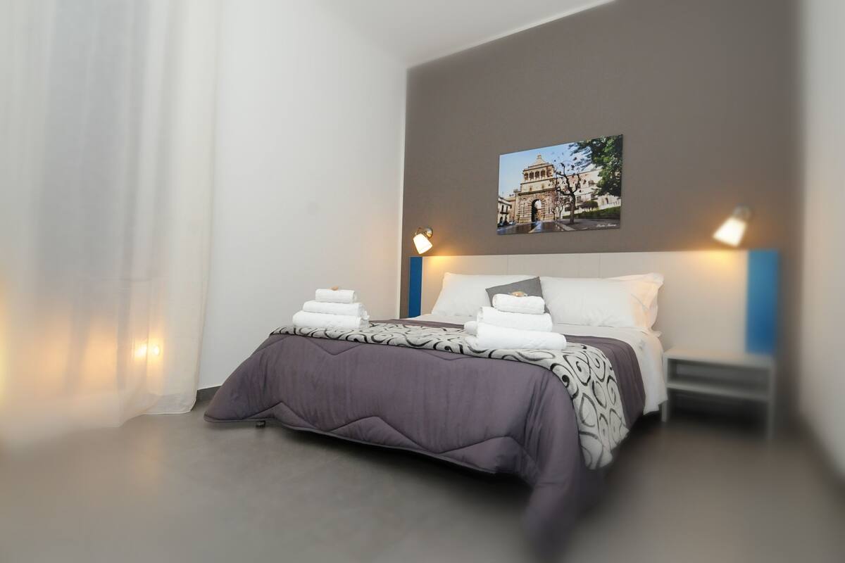 Cama bem arrumada com colcha estampada e almofadas, um quadro na parede e abajures laterais.