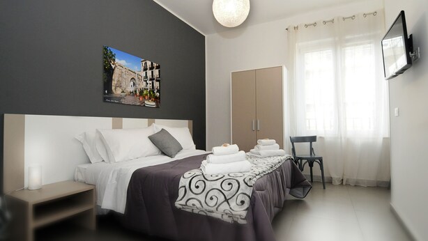 Een moderne hotelkamer met een groot bed, een flatscreen-televisie, een schilderij van een straatszene en uitzicht op het groen door dunne gordijnen.