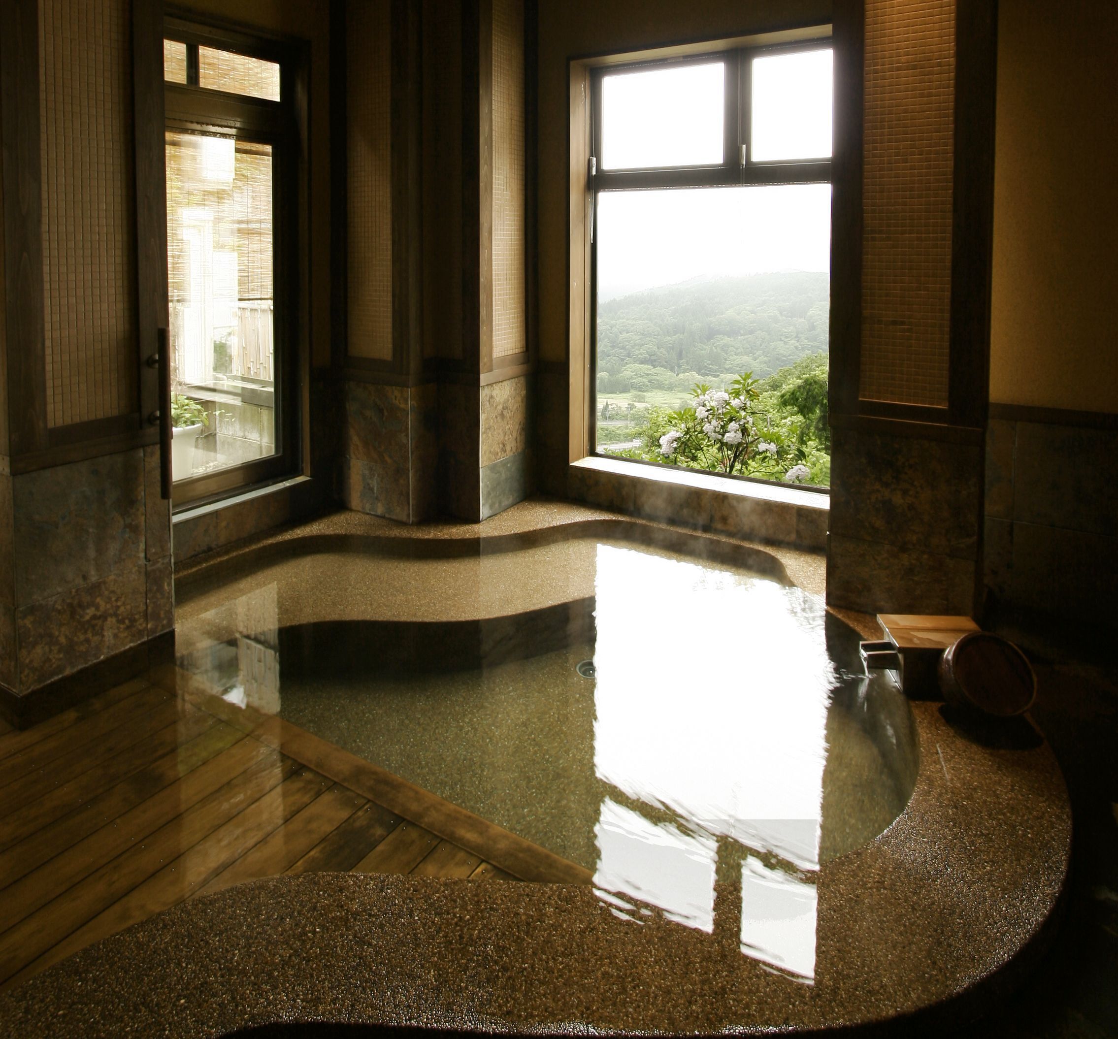 A spa room with a large, circular hot tub, a view of greenery outside, and a wooden floor.