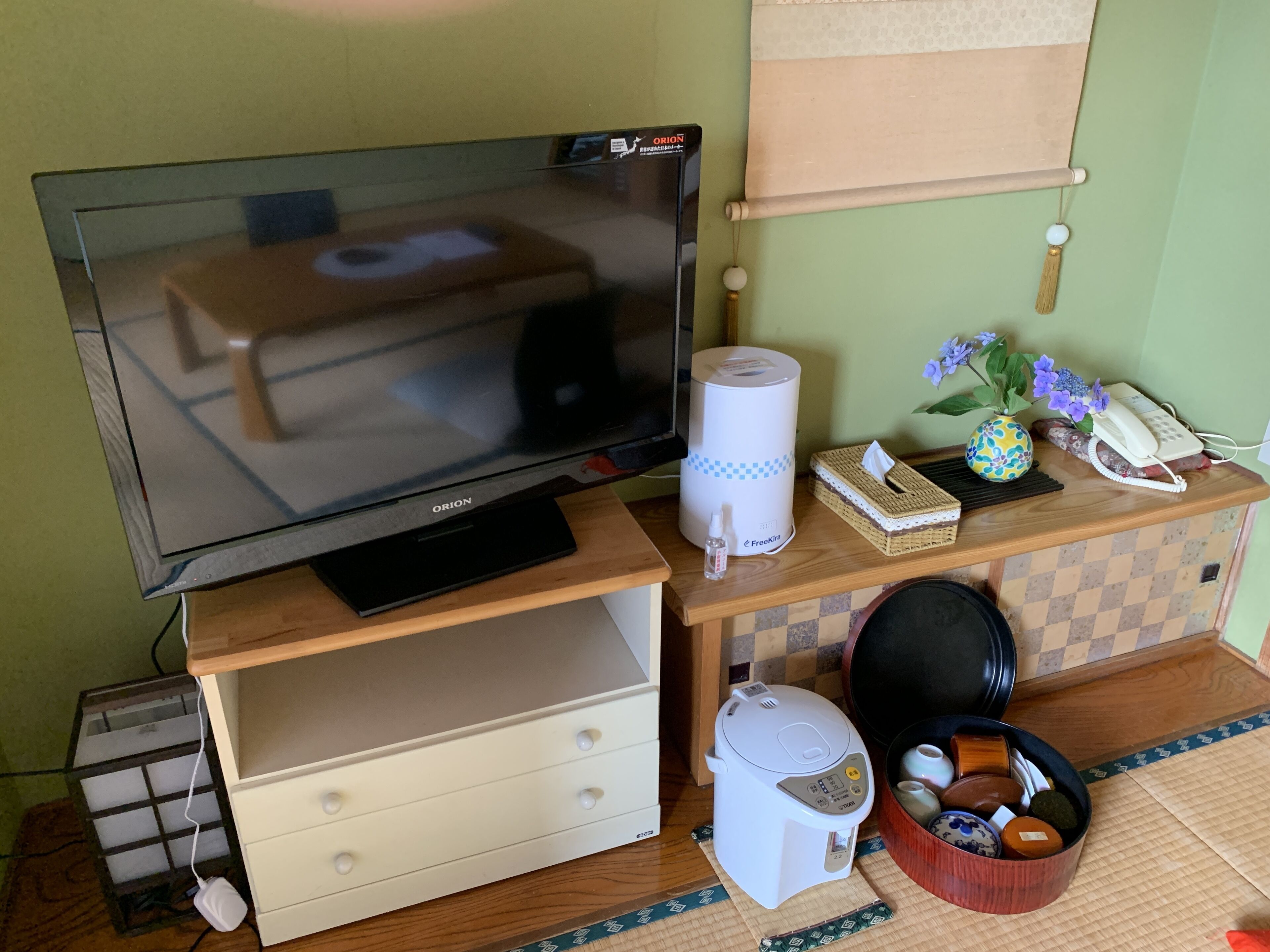 A room with a television on a wooden stand, a white air purifier, a small table with a tissue box, a phone, and a decorative vase with flowers.