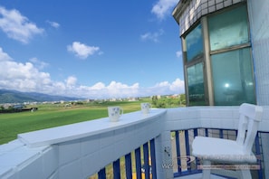Een balkon met uitzicht op een groen veld en bergen, een witte balustrade en twee witte stoelen.