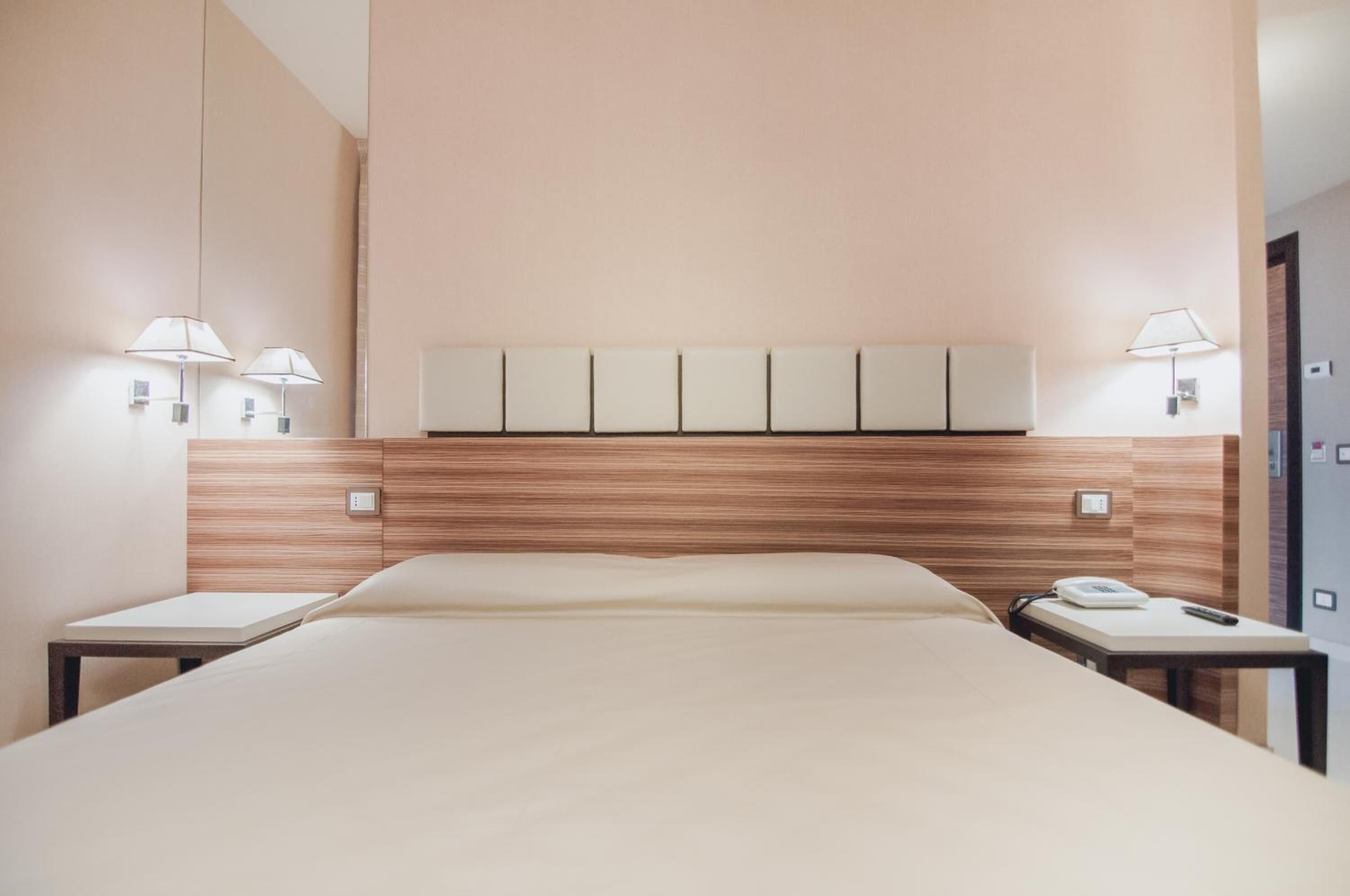 A hotel room with a large bed, bedside tables, a wooden headboard, wall-mounted lamps, and a telephone.