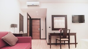 Een hotelkamer met een rode bank, een houten deur, een bureau met stoel, een aan de muur bevestigde televisie en een airconditioning.
