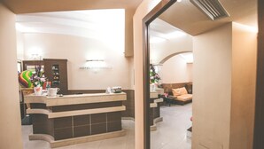 Een hotel receptie met een receptiebalie, een bord aan de muur, een zithoek met een bank en een sierplant.