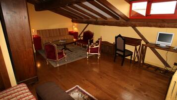 A loft-style room with exposed wooden beams, a red-framed window, a sofa, two armchairs, a small table, and a laptop on a wooden desk.