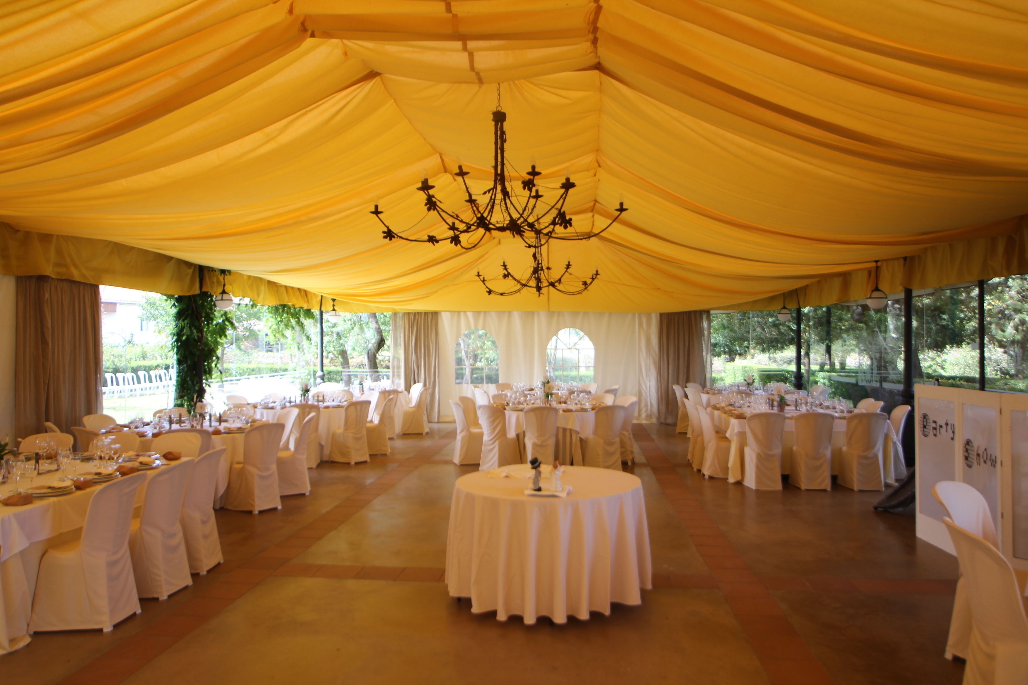 Un espacio para eventos con carpas, mesas redondas dispuestas para un encuentro formal, con una lámpara de araña central e iluminación natural gracias a grandes ventanales.